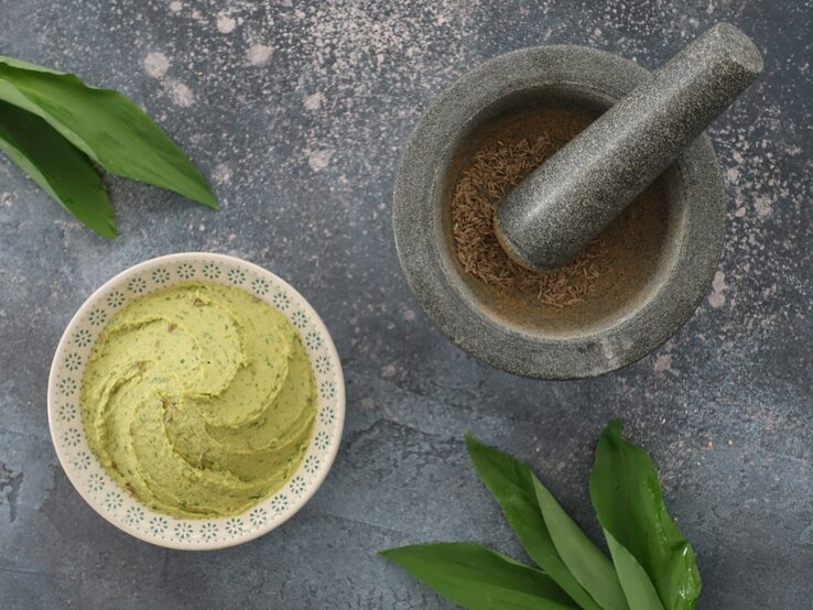 Eine Schale Bärlauch-Hummus in der Draufsicht, daneben ein Mörser mit Pfeffer und einige Bärlauch-Blätter.