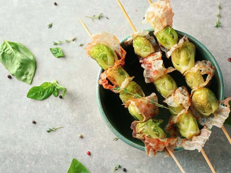 Drei Spieße mit Rosenkohl und Speck in einer Schüssel auf hellem Untergrund umgeben von Kräutern.