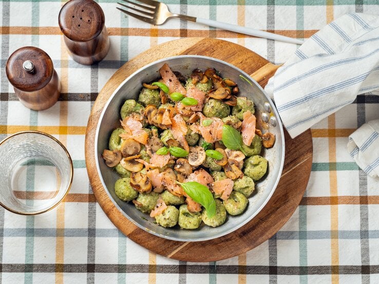 Draufsicht: Auf einem karierten Stoff stehen ein Salz- und ein Pfefferstreuer, ein Trinkglas, eine Gabel und ein rundes Holzbrettchen. Auf dem Brettchen steht eine Metallpfanne mit Bärlauch-Gnocchi mit Lachs.