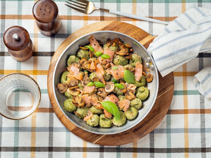 Draufsicht: Auf einem karierten Stoff stehen ein Salz- und ein Pfefferstreuer, ein Trinkglas, eine Gabel und ein rundes Holzbrettchen. Auf dem Brettchen steht eine Metallpfanne mit Bärlauch-Gnocchi mit Lachs.