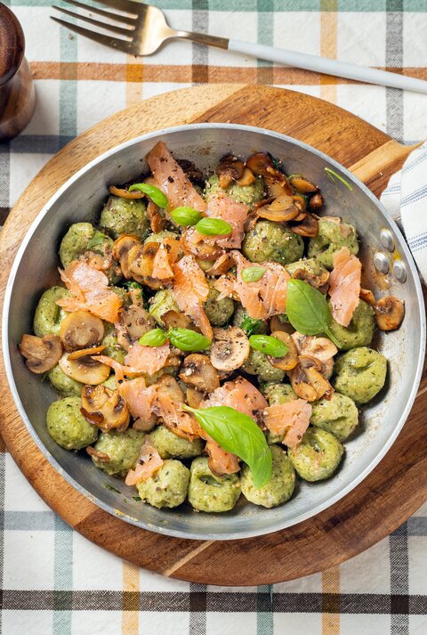 Draufsicht: Auf einem karierten Stoff stehen ein Salz- und ein Pfefferstreuer, ein Trinkglas, eine Gabel und ein rundes Holzbrettchen. Auf dem Brettchen steht eine Metallpfanne mit Bärlauch-Gnocchi mit Lachs.