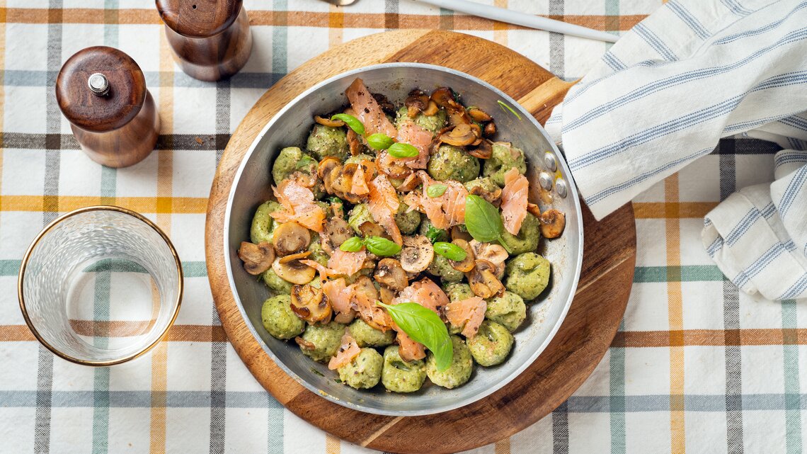 Draufsicht: Auf einem karierten Stoff stehen ein Salz- und ein Pfefferstreuer, ein Trinkglas, eine Gabel und ein rundes Holzbrettchen. Auf dem Brettchen steht eine Metallpfanne mit Bärlauch-Gnocchi mit Lachs.