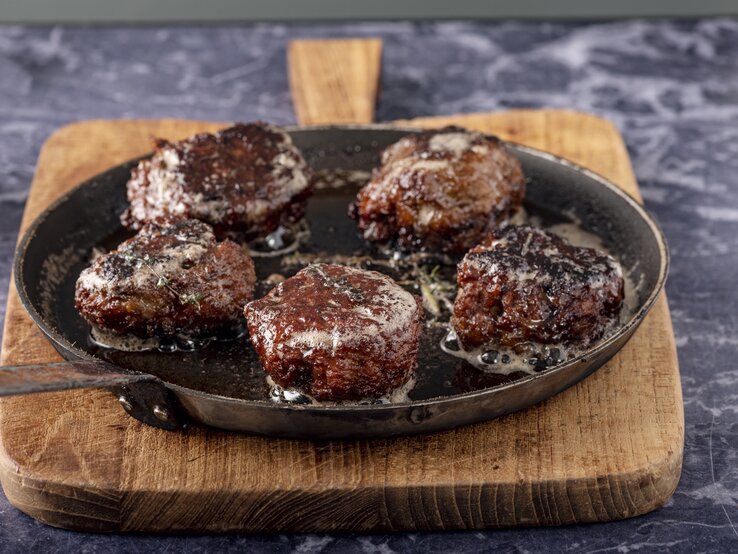 Auf einer Marmorplatte liegt ein dickes Holzbrettchen. Auf dem Holzbrettchen ist eine gusseiseren Pfanne. In der Pfanne sind frisch gebratene, knusprige Wildpflanzerl.