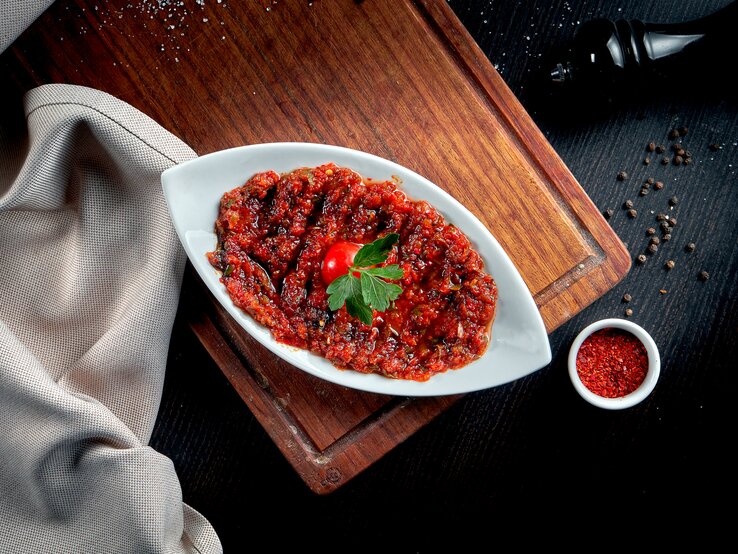 Draufsicht: Auf einem dunklen Untergrund liegt ein Holzbrett. Auf dem ist eine weiße Schale mit Acili Ezme, einer scharfen türkischen Gewürzpaste. Daneben liegen ein Tuch, eine Pfeffermühle sowie eine Schale mit einem roten Gewürzpulver.