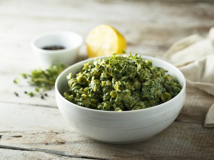 Eine weiße Schale Spirelli mit Rosenkohl und Tofu, garniert mit frischem Oregano. Im Hintergrund unscharf eine halbe Zitrone und eine kleine Schale sowie einige Kräuter.