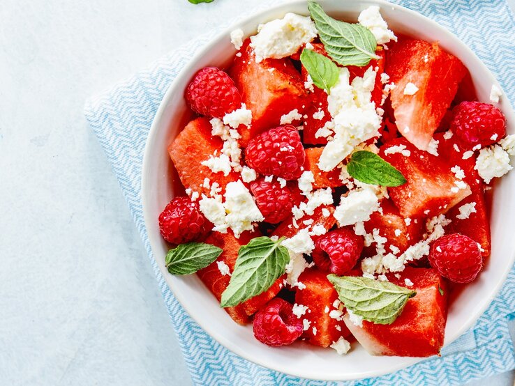 Wassermelonensalat-mit-Feta Wassermelonenslata mit Minze, Himbeeren und Feta in einer weißen Schale auf einem blauen Tuch. Auf einem hellen Tisch mit zwei Holzgabeln an der rechten Seite.