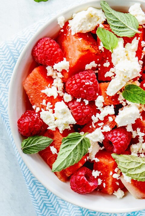 Wassermelonenslata mit Minze, Himbeeren und Feta in einer weißen Schale auf einem blauen Tuch. Auf einem hellen Tisch mit zwei Holzgabeln an der rechten Seite.