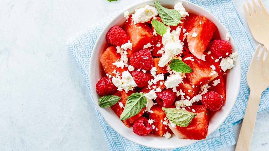 Wassermelonenslata mit Minze, Himbeeren und Feta in einer weißen Schale auf einem blauen Tuch. Auf einem hellen Tisch mit zwei Holzgabeln an der rechten Seite.