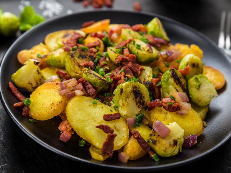 Gröstl mit Rosenkohl Ein schwarzer Teller mit Gröstl mit Rosenkohl auf einem schwarzen Tisch.