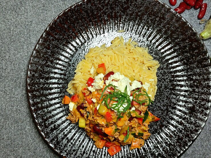 Reisnudeln auf schwarzem Teller und grauem Untergrund, mit mediterranem Gemüse an der Seite.