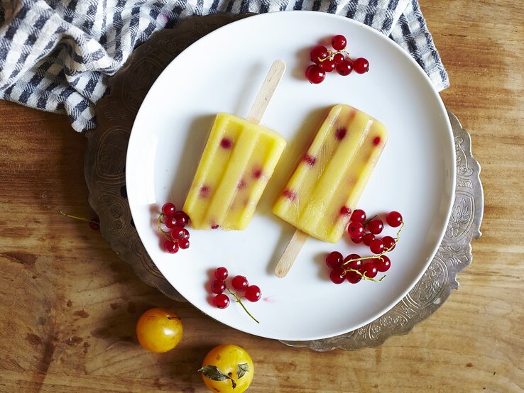 Zwei selbstgemachte Mirabellen-Preiselbeer Eis am Stiel auf einem weißen Teller mit frischen Preiselbeeren und Mirabellen umgeben, von oben vor einem Holzhintergrund.