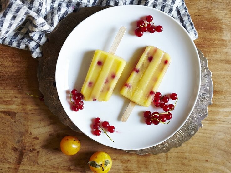 Zwei selbstgemachte Mirabellen-Preiselbeer Eis am Stiel auf einem weißen Teller mit frischen Preiselbeeren und Mirabellen umgeben, von oben vor einem Holzhintergrund.