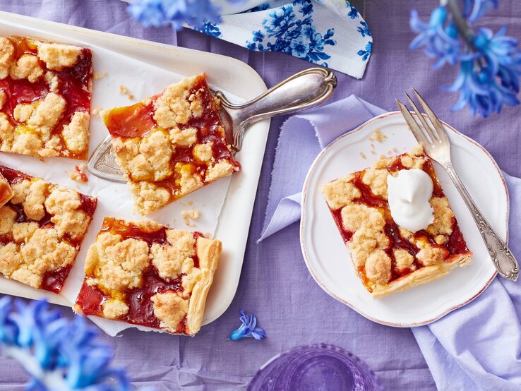 Ein weißes Kuchenblech mit Stücken des Prasselkuchens mit Erbeeren und Mango darauf neben einem Teller mit einem weiterem Stück, einer Gabel und blauen Blumen, vor einem fliederfarbenen Hintergrund, von oben. 