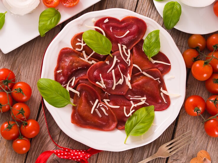 Einige rote Herz-Ravioli aus Rote-Bete-Saft und Tomatenmark mit Hackfleischfüllung liegen auf einem weißen Teller. Die Nudeln sind mit Streukäse und Basilikum angerichtet.
