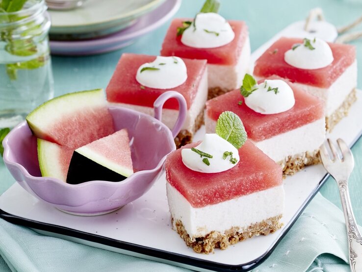 Diese Wassermelonen-Kokos-Schnitten schmecken genau so süß, wie sie aussehen. Noch dazu sind sie erfrischend und schnell gemacht! Auf einem Tablett liegen mehrere Wassermelonen-Kokos-Schnitten. Sie sind mit Minze verfeinert.