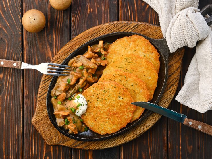Haferpuffer-Pfifferlingsrahm Eine gusseiserne Pfanne mit den Haferpuffern an einem Pfifferlingsrahm von oben fotografiert auf einem dunklen, rustikalen Holztisch. Außerdem sind auch Messer, Gabel und ein helles Tuch mit auf dem Bild.