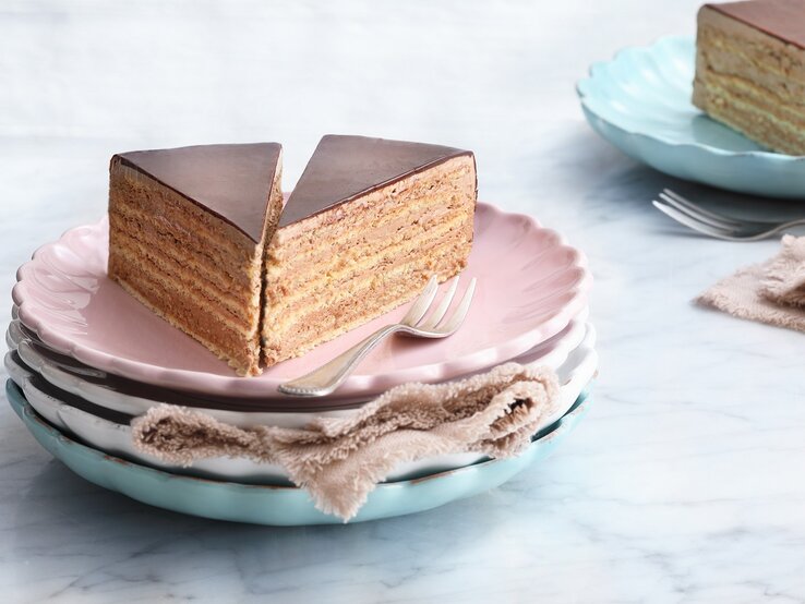 Auf einem Teller sind zwei Stück  Prinzregententorte serviert. Sie ist mit einer dünnen Schokoladenschicht überzogen. 