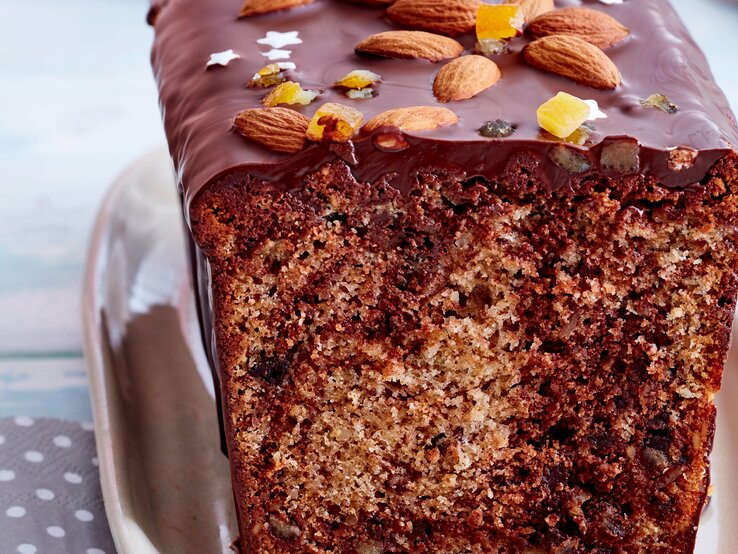 Angeschnittener Marmor-Gewürzkuchen mit Schokoladen-Glasur, Mandeln, Zuckersternen und Orangeat auf rosafarbenem Teller, weiß gepunkteter grauer Serviette und hellblauem Tischtuch in der Frontalsicht.