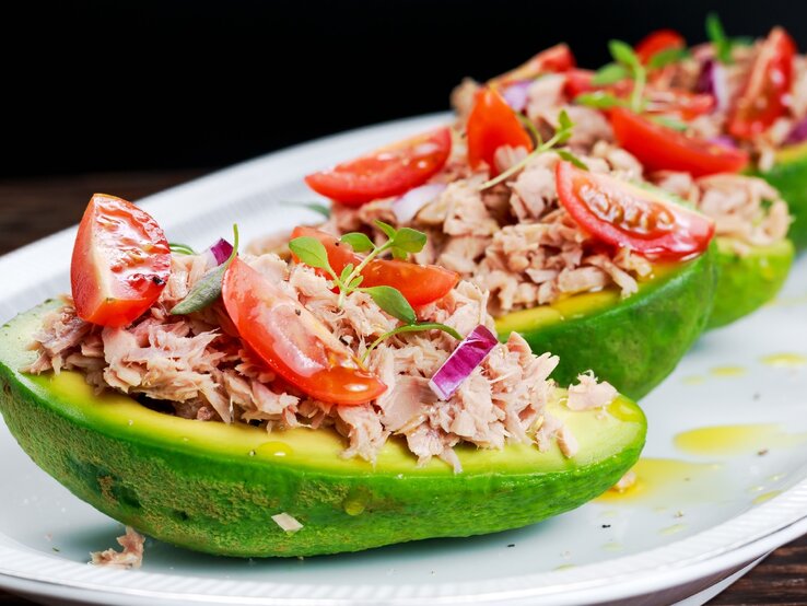 Ein weißer länglicher Teller mit vier Avocadohälften, in denen der Thunfischsalat angerichtet ist vor schwarzem Hintergrund.