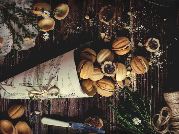 Eine Papiertüte aus Notenblättern gefüllt mit vielen Stücken der polnischen Zaubernüsse Orzeszki, einige im ganzen Stück, andere sind geöffnet, sodass man die Schokoladenfüllung sehen kann. daneben sind weitere Notenblätter, Blumen und ein Messer zu sehen, vor einem Holzhintergrund von oben. 