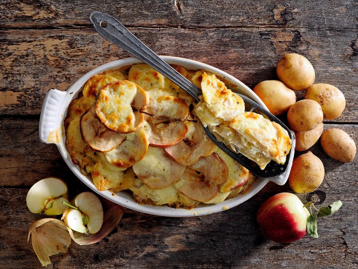 Mit Käse überbackener Kartoffel-Apfel-Auflauf in weißer Auflaufform auf hölzernem Untergrund. Darum herum sind Kartoffeln und Äpfel verteilt. 