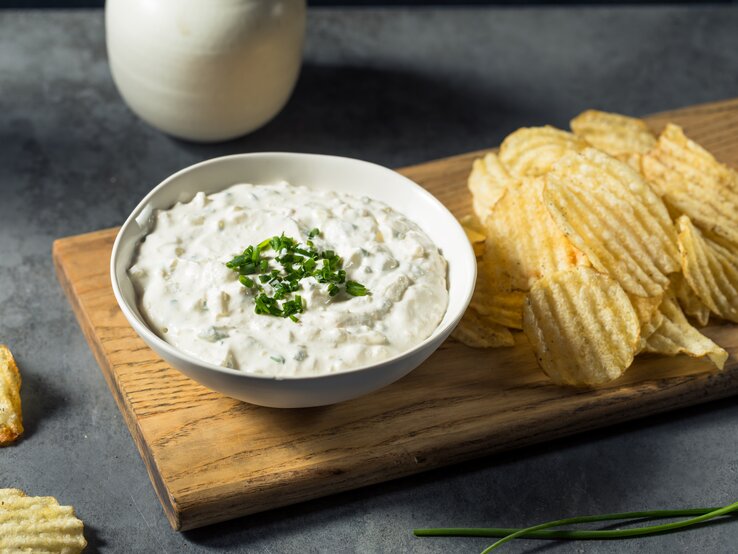 Französischer Zwiebeldip in weißer Schale auf Holzbrett auf dunkelgrauem Tisch. Auf dem Brett neben dem Dipschälchen geriffelte Chips. Am oberen Bildrand ein Kännchen mit Sahne. Gräser und Chips als Deko in Bild. Frontalansicht.
