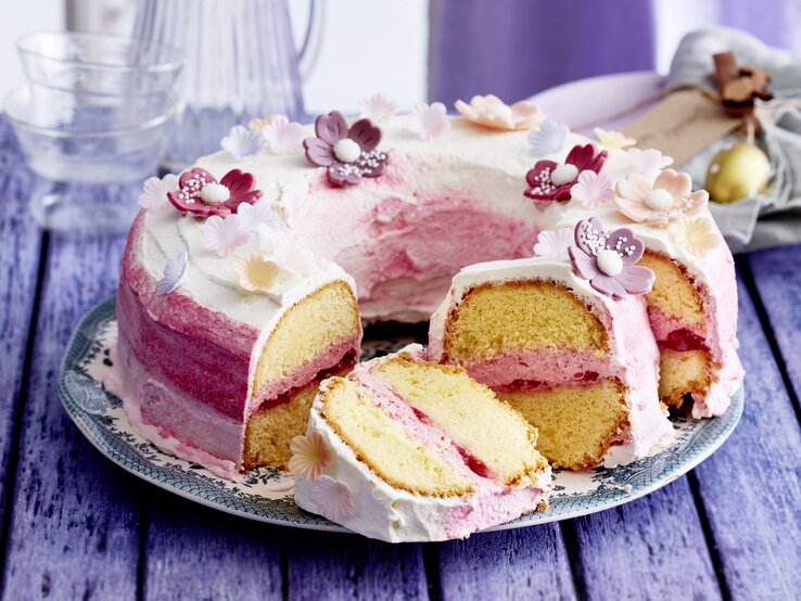 Auf einem Teller liegt ein Blütenkranz-Kuchen. Er ist mit Sahne eingestrichen und mit essbaren Blüten dekoriert. Man erkennt die Erbdeerfüllung in der Mitte.