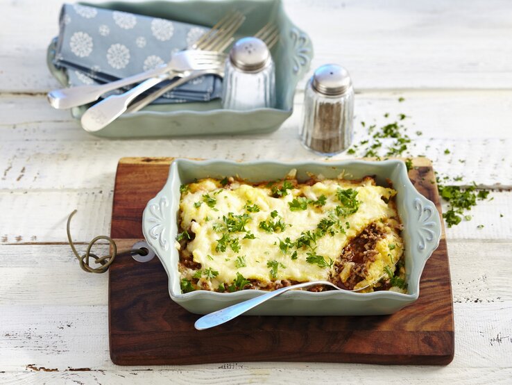 Eine Auflaufform mit Polenta-Hackfleischgratin. Daneben weiteres Besteck und Geschirr und Salz und Pfeffer. Das Ganze auf einem hölzernen Untergrund. 