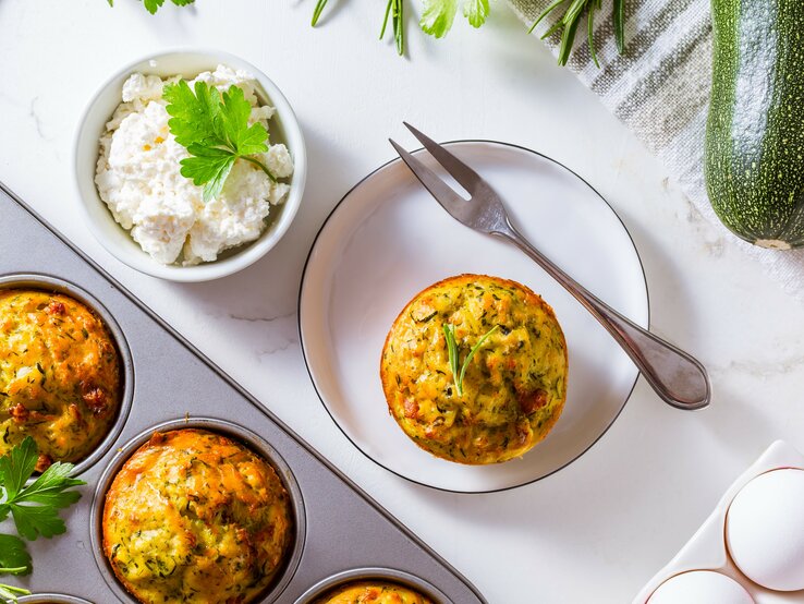 Die Muffin-Form mit den Muffins auf einem Tisch mit weißer Tischdecke. Daneben weiße Eier und ein Teller mit einem Muffin aus der Form.