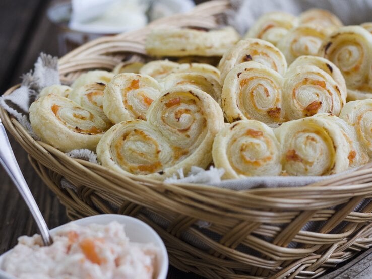 Blätterteigschnecken mit Lachs und Meerettich-Frischkäse, gestapelt in einem großen Korb, serviert mit der Lachs-Creme auf einem hölzernen Tisch. 
