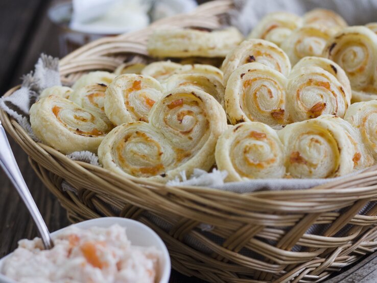 Blätterteigschnecken mit Lachs und Meerettich-Frischkäse, gestapelt in einem großen Korb, serviert mit der Lachs-Creme auf einem hölzernen Tisch. 