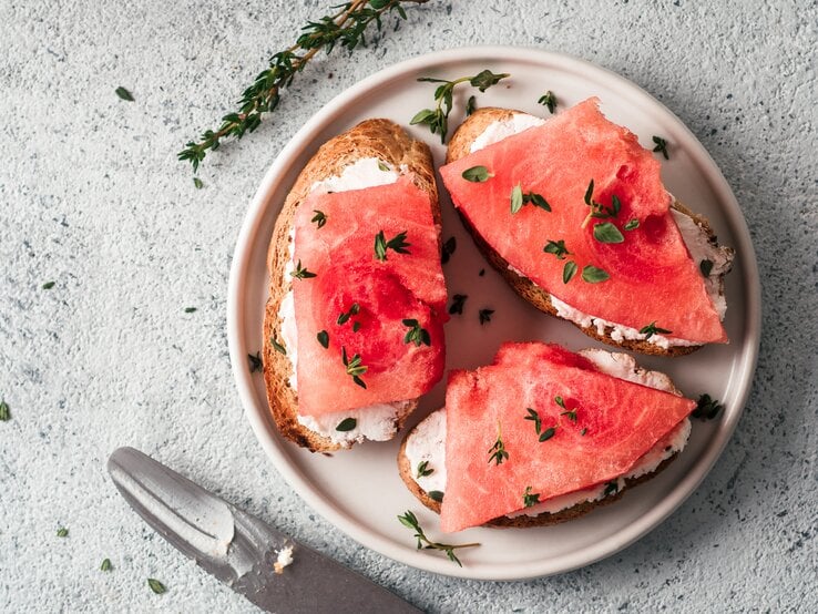 Ein Teller mit Brotscheiben, auf denen Frischkäse und veganer Thunfisch aus Wassermelone liegt. Bestreut mit frischen Kräutern. Draufsicht.