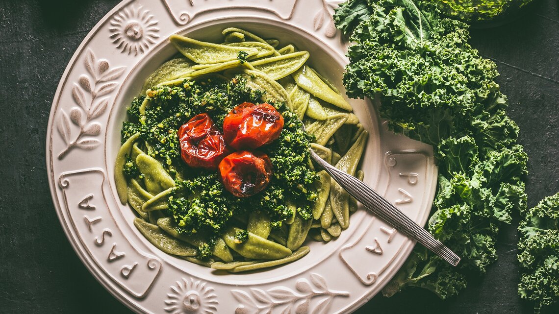Ein Teller Grünkohl-Pasta mit Zitrone und Tomaten in der Draufsicht, daneben frischer Grünkohl und ganze Tomaten.