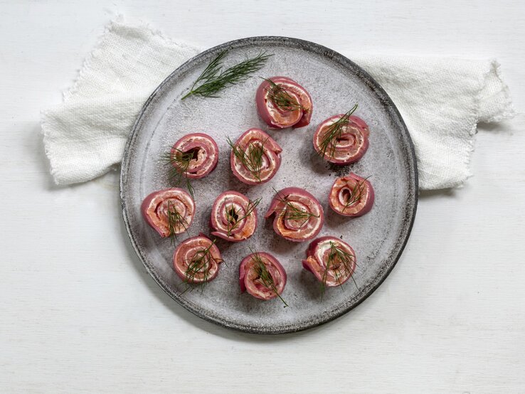 Rote-Bete-Pfannkuchenröllchen mit Lachs und Meerrettich, gerollt und in kleine Happen geschnitten serviert auf einem grauen Tisch auf einem weißen Tisch.