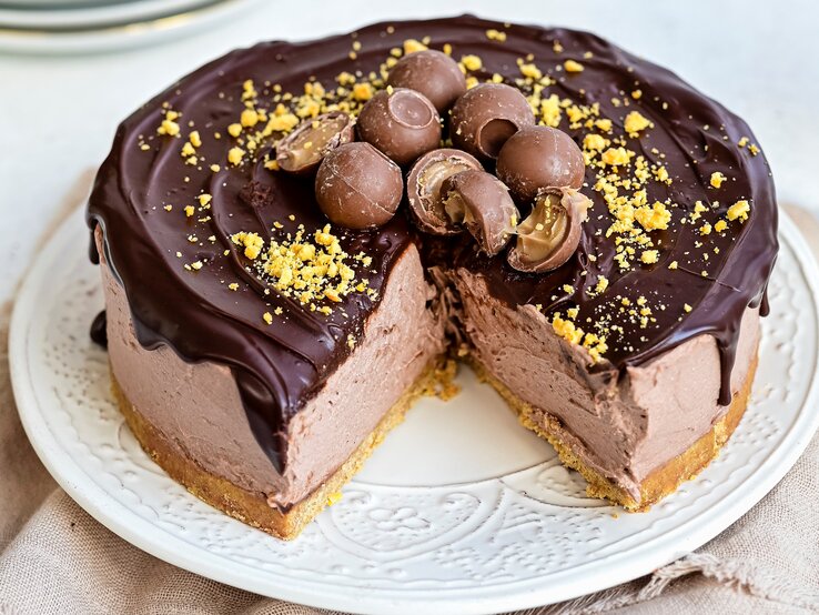 Ein weißer Teller mit einem angeschnittenen Baileys-Käsekuchen mit Schokoglasur auf einem braunen Tischtuch vor einem hellen Hintergrund.  | © StockFood / Parissi, Lucy