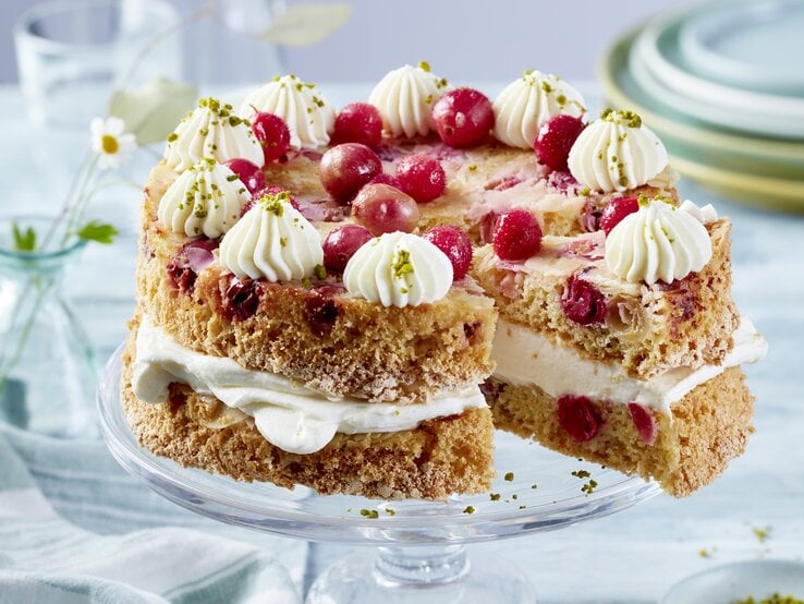 Eine zweilagige Torte mit hellem Teig und roten Johannisbeeren, gefüllt und dekoriert mit weißer Sahnecreme. Die Torte ist auf einem Glas-Tortenständer angerichtet und mit weiteren Johannisbeeren und gehackten Pistazien garniert. Im Hintergrund stehen gestapelte Teller und Glasvasen mit kleinen Blumen.