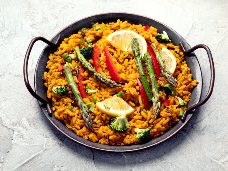Eine vegane Paella mit grünem Spargel, Zitronenscheiben, Paprika und Brokkoli in einer typischen Paella-Pfanne.