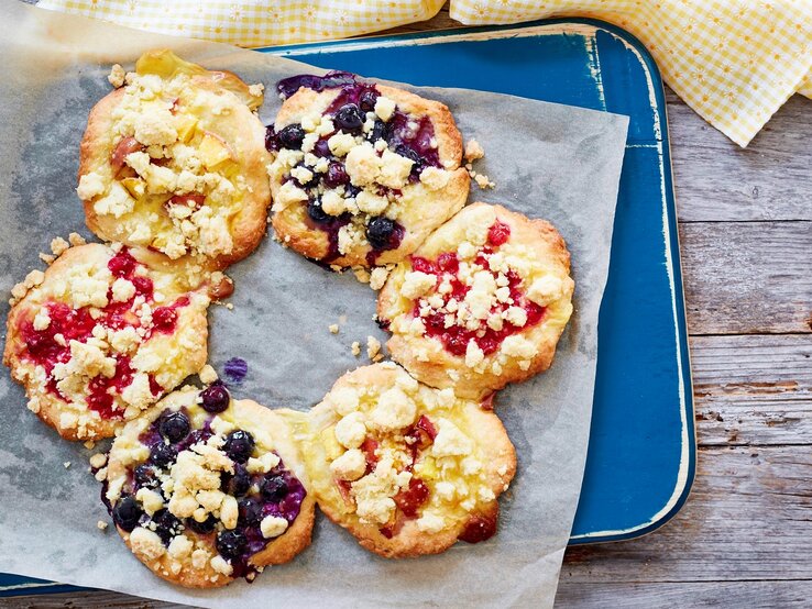 Streuseltaler-Kranz mit fruchtigen Beeren und Vanillepudding, serviert auf einem hellblauen Tablett auf einem groben Holztisch.