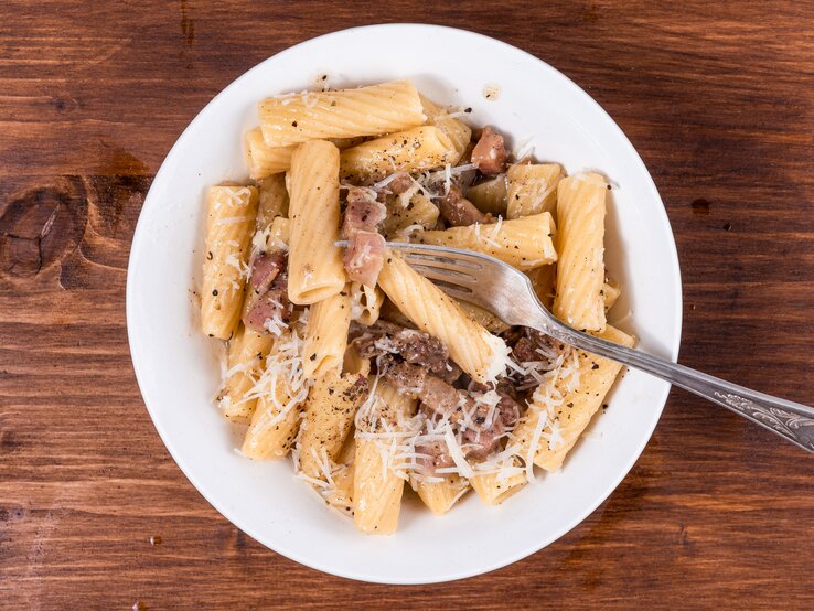 Pasta alla gricia Rigatoni mit gebratenem Guanciale und Pecorino auf weißem Teller. Dieser steht auf einem Holzuntergrund.