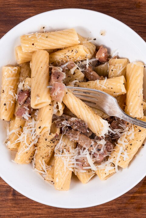 Rigatoni mit gebratenem Guanciale und Pecorino auf weißem Teller. Dieser steht auf einem Holzuntergrund. 
