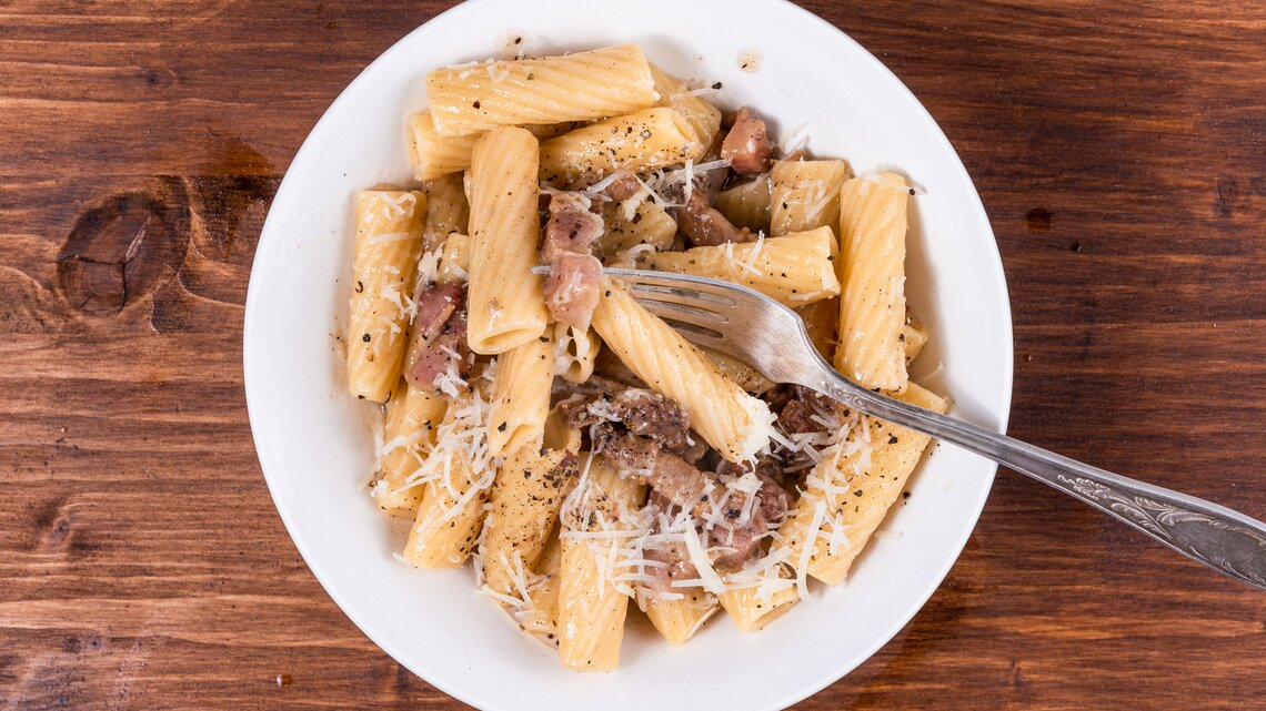 Rigatoni mit gebratenem Guanciale und Pecorino auf weißem Teller. Dieser steht auf einem Holzuntergrund. 