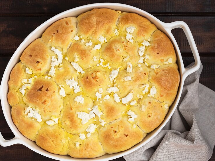 Eine weiße Auflaufform mit dem Schafskäsebrot auf einem dunklen Holztisch mit grauer Tischdecke.