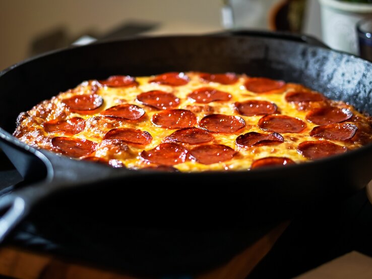 Pizza mit Salami in einer gusseisernen Pfanne. Dahinter verschiedene Behälter. 