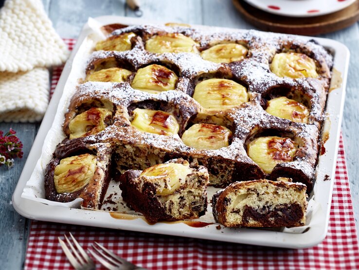 Apfel-Stracciatella-Kuchen Ein Apfel-Stracciatella-Kuchen mit Gelee und Puderzucker in einem tiefen Blech. Der Kuchen ist angeschnitten und zwei Stücken liegen am Rand der Form.