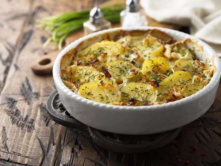 Ein Carbonara-Kartoffelauflauf in einer runden Auflaufform auf einem Holztisch, im Hintergrund unscharf Salz- und Pfefferstreuer, Kräuter, ein Brett und ein Geschirrtuch.