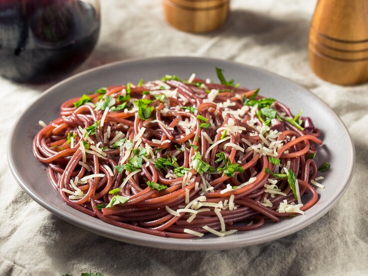 Auf einem Tisch steht ein Teller mit Rotwein-Spaghetti. Daneben sind halb zu sehen: Eine Karaffe mit Rotwein sowie ein Salz- und Pfefferstreuer.