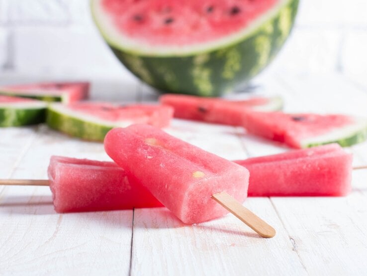 Ein Aperol Eis am Stiel, Stücke einer Wassermelone und eine halbierte Wasserlemone auf einem Untergrund aus Holz.