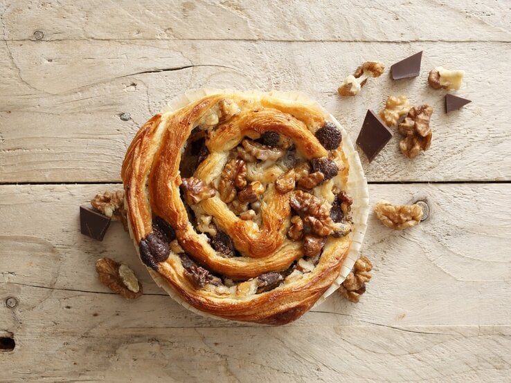 Auf einer Arbeitsplatte aus Holz liegt eine der Blätterteigschnecken mit Schokolade und Walnüssen. Sie ist golbraun gebacken und mit Walnüssen und Schokolade belegt. 