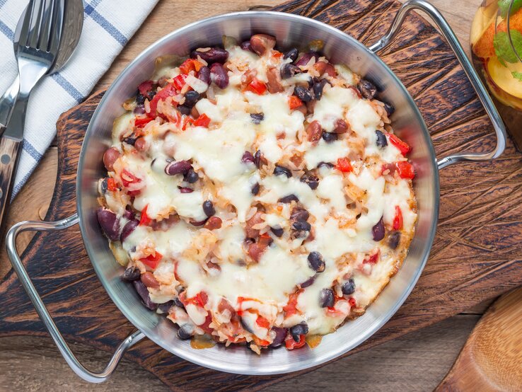 Silberne Form gefüllt mit Paprika-Reis-Auflauf mit Bohnen, angerichtet auf einem Holbrett. Das Szenario ist dekoriert mit einem Geschirrtuck, Besteck und einem Glas Limonade. 