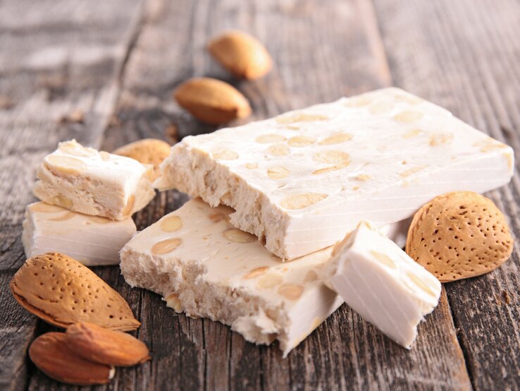 Stücke des italienischen Nougats Terrone di Cremona auf einem Holztisch neben Mandeln und Mandelkernen. 